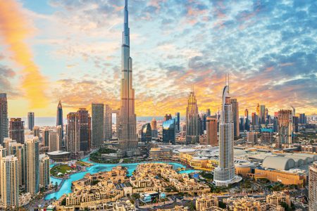 View of the skyline of Dubai with the iconic Burj Khalifa towering over the city. The sky is clear and blue with a few clouds. In the foreground is a body of water with boats floating on it